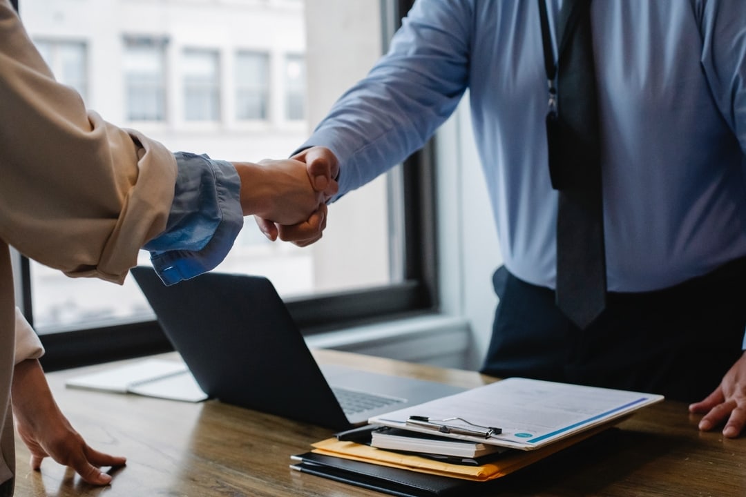 Two professionals shaking hands over a desk with a laptop and documents