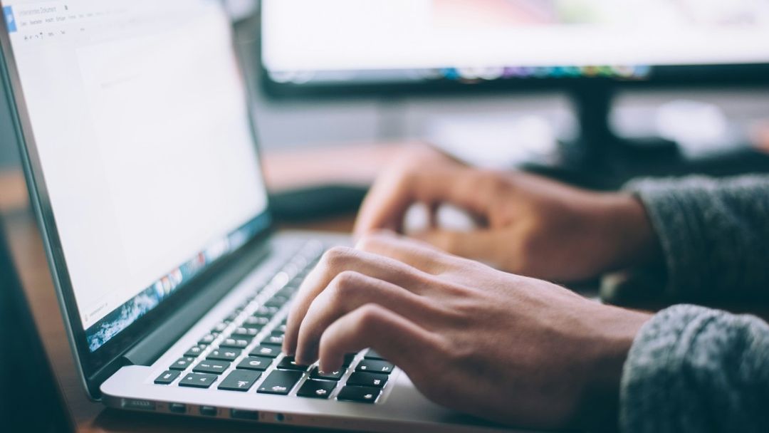 Close up of hands typing a document on a laptop beside a second monitor