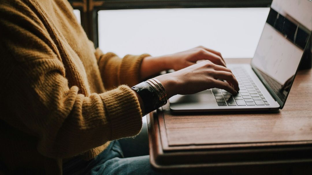 Person in a sweater typing on a laptop at a wooden desk